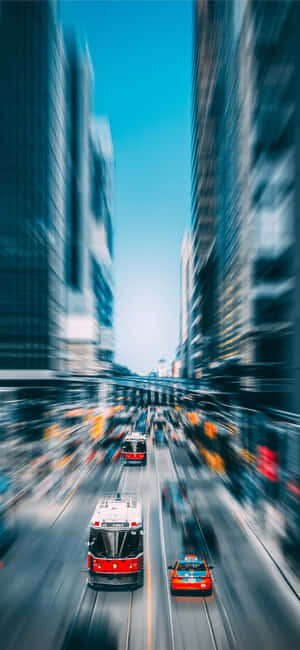 紅色電車和汽車在藍天下高樓林立的城市街道上模糊掠過。 — Moto E13 壁紙
