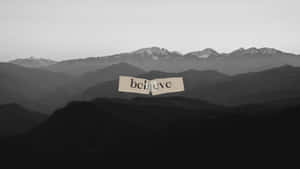 撕破的紙上寫著believe，漂浮在灰階山脈之上。 — 黑白美學 壁紙