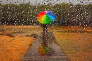 人拿著彩虹傘在濕路上行走，透過有雨痕的窗戶。 — 雨天 壁紙