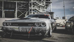 一輛骯髒的黑色 Dodge Challenger SRT 停在賽道上，背景是工業用鷹架。 — 酷車 壁紙