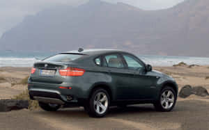綠色BMW X6 SUV停泊在近海的沙灘上，遠方有山脈。 — Steam Deck OLED 壁紙