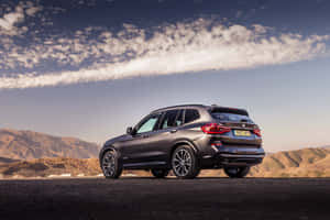 深灰色BMW X3 SUV停泊在沙漠公路上，背景有山脈和日落天空。 — Surface Pro 壁紙