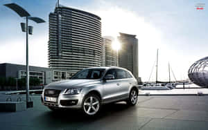 銀色 Audi Q7 SUV 停泊在海濱附近，有帆船和現代建築。 — Steam Deck OLED 壁紙