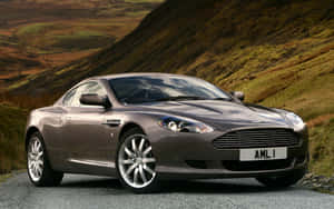 銀色Aston Martin DB9跑車停泊在碎石路上，靠近綠色山丘。 — Steam Deck OLED 壁紙