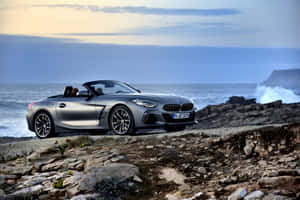 銀色BMW Z4開篷跑車停在岩石懸崖上，下方海浪在黃昏時分拍打。 — Surface Pro 壁紙
