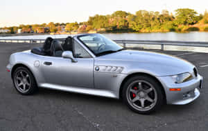 銀色BMW Z8開篷車停泊在水邊附近，背景有樹木和日落。 — Steam Deck OLED 壁紙