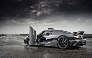 銀色Koenigsegg Agera在空路上打開剪刀門，背景是暴風雨的天空。 — 黑白 壁紙