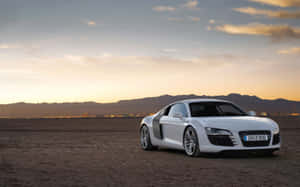 夕陽下沙漠中的白色奧迪R8跑車，背景有城市燈光和山脈。 — Audi R8 GIF 壁紙