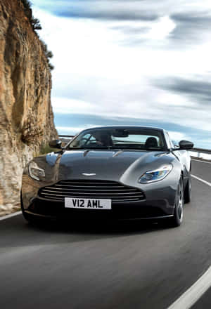 銀色Aston Martin V12開篷跑車在懸崖附近的海岸公路上飛馳。 — iPad (A16) 壁紙