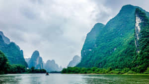 一艘小船漂浮在平靜的河流上，周圍環繞著高聳的綠色喀斯特山脈，天空多雲。 — iMac 壁紙