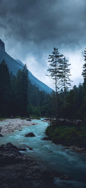 一條快速流動的河流在暴風雨的天空下蜿蜒穿過茂密的森林，遠處有山脈。 — iPhone 17e 壁紙