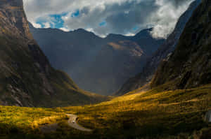 蜿蜒的道路穿過一個山谷，上方有金色陽光和暴風雲。 — MacBook Air M3 壁紙
