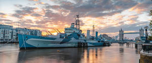 藍色軍艦停泊在河邊，背景是倫敦塔橋和日落時分的城市天際線。