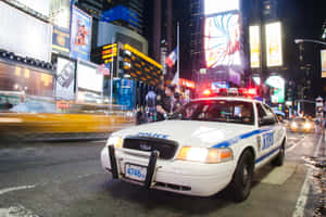 NYPD警車在夜晚停在繁忙的時代廣場街道上，周圍是明亮的廣告牌和車流。 — Surface Pro 壁紙
