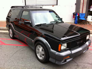 黑色GMC Safari SUV停放在鋪砌的空地上，附近有紅線和椅子。 — 4:3 壁紙