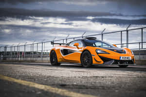 橙色McLaren跑車在多雲天空下的賽道上。 — Surface Pro 壁紙