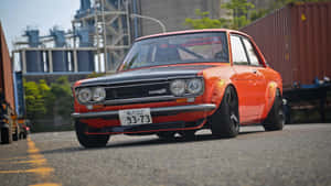 橙色和黑色經典Datsun跑車停在工業集裝箱附近。 — 酷車 壁紙