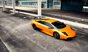 橙色林寶堅尼 Murciélago 停泊在火車軌道和工業建築物附近的混凝土上。 — 蘭博基尼 壁紙