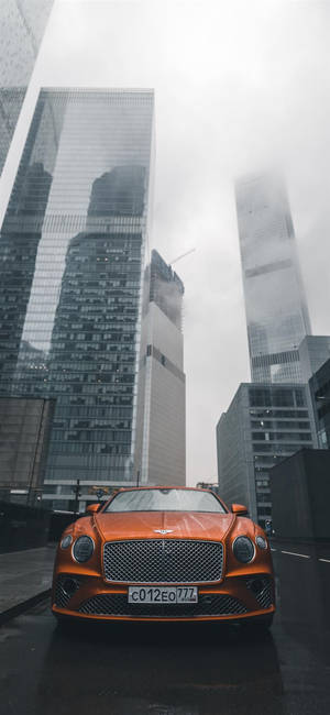橙色賓利豪華車停在濕漉漉的城市街道上，背景是霧氣中高聳的玻璃建築。 — iPhone 13 Mini 壁紙
