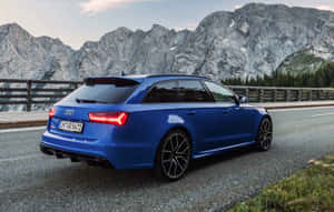 藍色 Audi RS6 Avant 在山路上行駛，背景有岩石山峰和樹木。 — 1280x800 壁紙