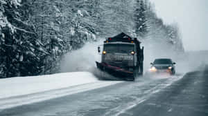 剷雪車清理道路上的積雪，一輛汽車在冬季天氣中跟在後面。 — Studio Display 壁紙