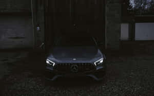 一架深灰色Mercedes-AMG在夜間停泊在碎石庭院中，車頭燈閃爍發光。