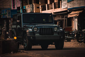 深綠色Mahindra Thar SUV停泊在塵土飛揚的街道上，伴隨著電單車和建築物。 — 啞光黑色 壁紙