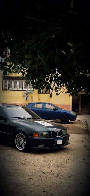 黑色BMW轎車帶白色條紋停在樹枝下，附近有藍色汽車和黃色建築物。 — Huawei 壁紙