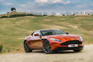 橙色Aston Martin跑車在托斯卡納山丘的土路上，夕陽時分。 — MacBook Air M2 壁紙