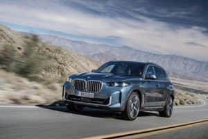 藍色BMW X5 SUV在沙漠公路上行駛，背景有山脈。 — Surface Pro 壁紙
