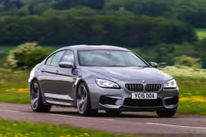 銀色BMW M6雙門跑車在鄉村道路上行駛，背景有綠色山丘和黃色花朵。 — Surface Laptop 壁紙