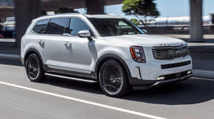 銀色Kia Telluride SUV在陽光明媚的城市高速公路上行駛，帶有動態模糊效果。 — 低調 壁紙