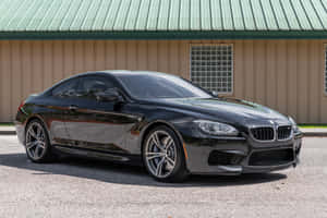 黑色BMW M6跑車停放在米色建築附近的碎石地上。 — Surface Laptop 壁紙
