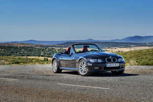 黑色BMW Z3開篷車配紅色座椅，停泊在山附近的碎石路上。 — Surface Laptop 壁紙