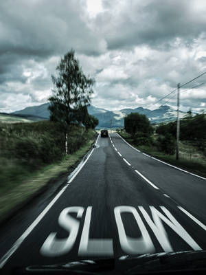 一輛汽車在塗有「SLOW」的道路上行駛，背景是陰雲密布的天空下的山巒。 — 道路 壁紙
