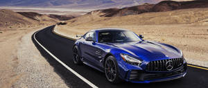 藍色 Mercedes-AMG GT 在沙漠公路上，背景有山脈。 — 藍色 壁紙