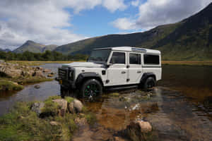 白色 Land Rover Defender 穿越一條有岩石河岸和背景綠色山丘的山溪。 — MacBook Air M2 壁紙