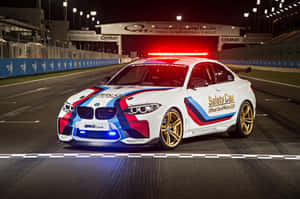 夜間賽道上帶有紅、藍、金色塗裝的白色BMW M2 Coupe安全車。 — Surface Pro 壁紙