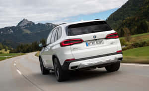 白色BMW X5 SUV在蜿蜒的山路上行駛，周圍有綠色的山丘和岩石山峰。 — Steam Deck OLED 壁紙