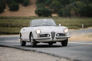白色復古Alfa Romeo Spider開篷車在日落時分行駛在鄉村道路上。 — Surface Pro 壁紙