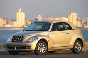 金色 PT Cruiser 敞篷車停在水邊，日落時分城市天際線。 — Surface Laptop 壁紙