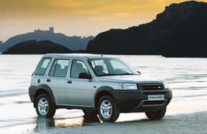 銀色 Land Rover Freelander 在日落時分停泊在岩石峭壁附近的濕沙灘上。 — MacBook Pro 14 壁紙