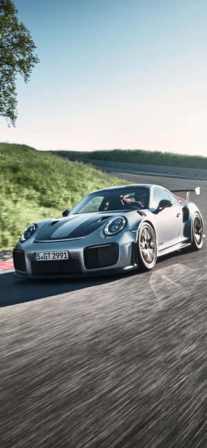 銀色保時捷 911 GT2 RS 在賽道上奔馳，背景有綠色山丘和藍天。 — iPhone 18 壁紙