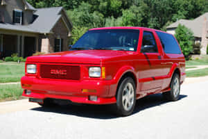 紅色GMC Typhoon SUV停放在住宅街道上，有房屋和綠化。 — 夏天 壁紙