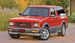 紅色GMC Typhoon SUV停放在磚砌車道上，後面有綠植。 — 2560x1440 壁紙