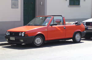 橙色復古Fiat 126 Spider開篷車停泊在陽光明媚的城市街道上。 — Surface Pro 壁紙