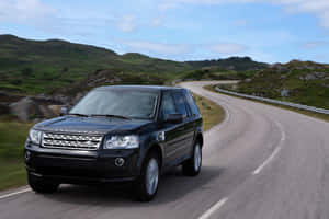 黑色Land Rover SUV在蜿蜒的山路上行駛，周圍是綠色的山丘和藍天。 — MacBook 壁紙