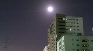 滿月在夜晚的城市建築上方發光，窗戶亮著燈。 — 月色夜空 壁紙