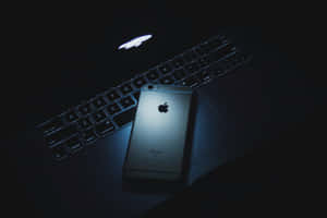 銀色 iPhone 6s 放在筆記型電腦鍵盤上，帶有發光的 Apple 標誌，在深藍色光線中。 — MacBook Pro M4 壁紙