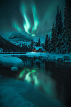 夜晚，綠色北極光倒映在河流中，一座被雪覆蓋的小屋。 — 日出 壁紙